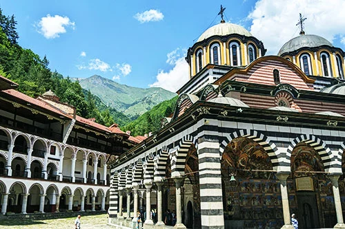Striped arches and painted domes of Rila Monastery below forested mountains, Bulgaria